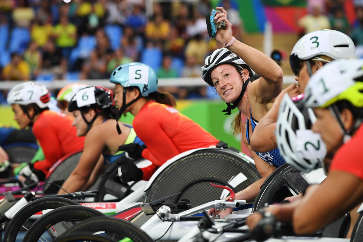 A woman in a racing wheelchair waving to the camera surrounded by other six female wheelchair racers