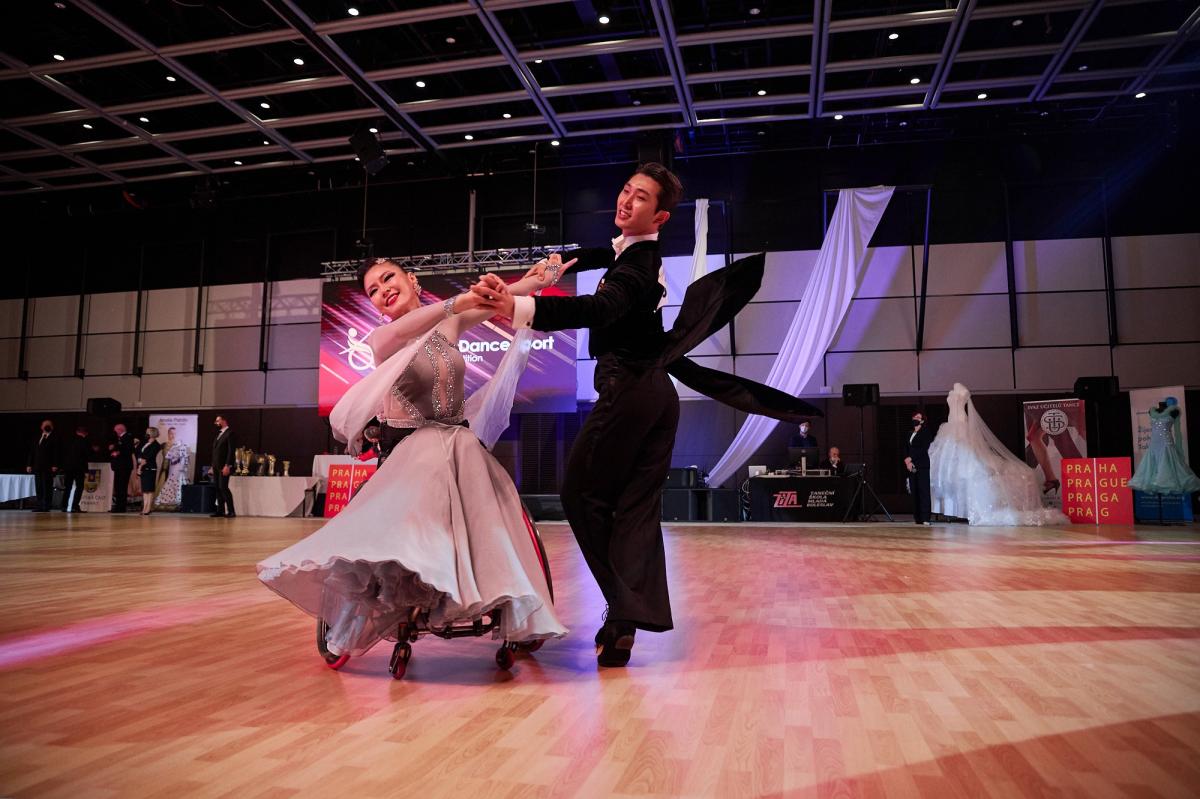 A female wheelchair dancer and her standing male partner in a Para dance sport competition observed by seven people standing in the background