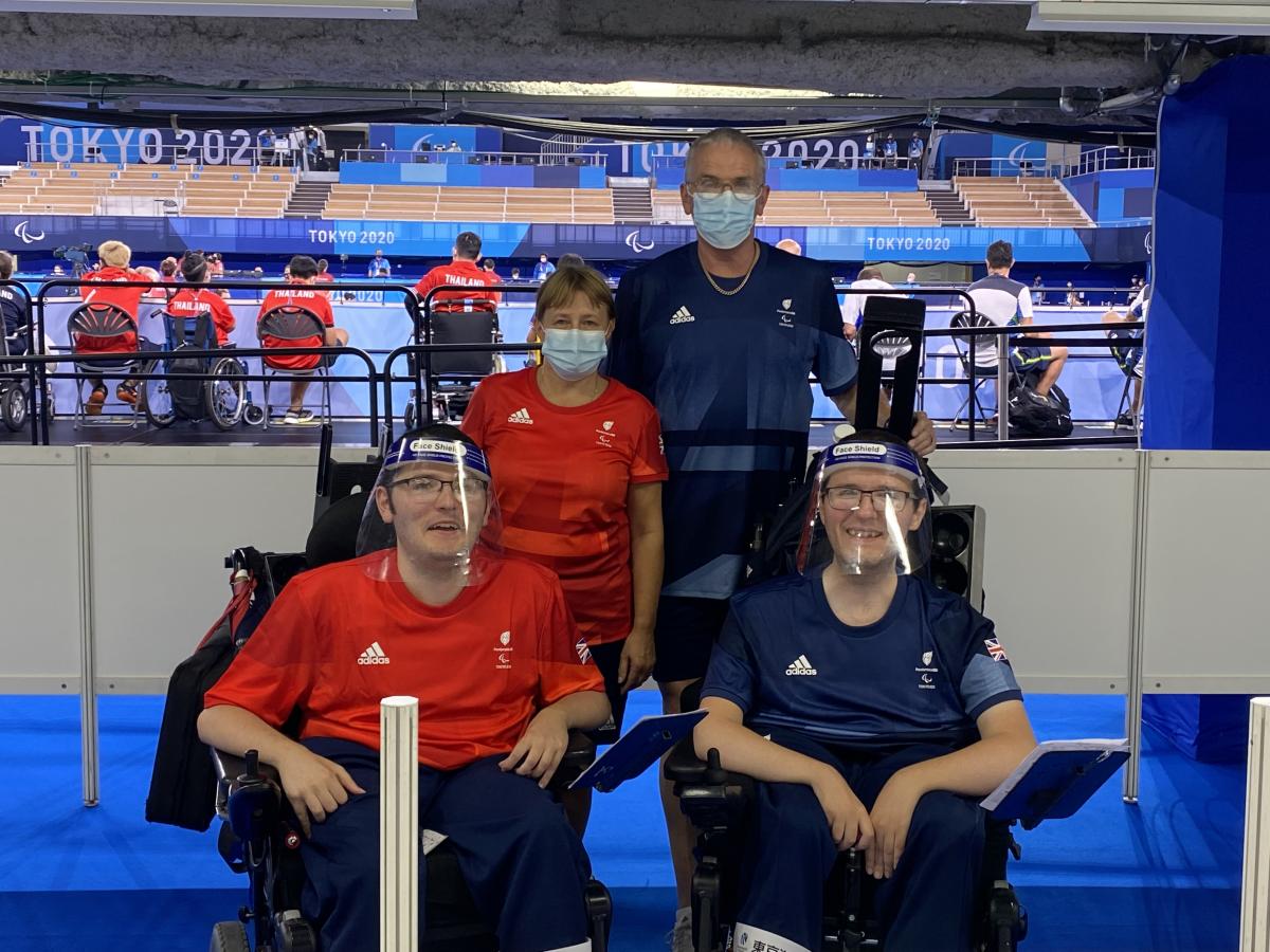 Linda stands behind Scott in his wheelchair, both wearing red, while Gary stands behind Jamie in his wheelchair, both in blue