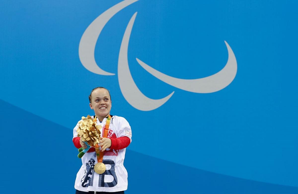 A short stature woman with her gold medal in front of a wall with the Paralympic Agitos
