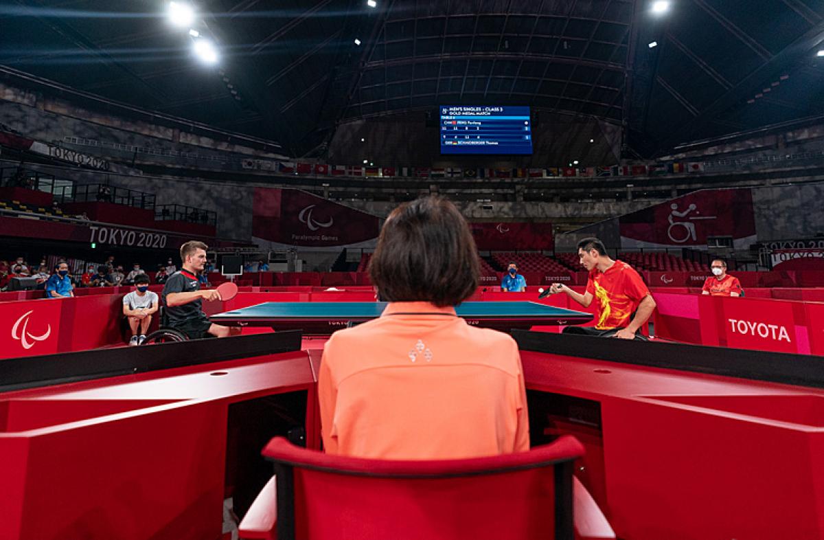Two male wheelchair table tennis players playing with judge looking on