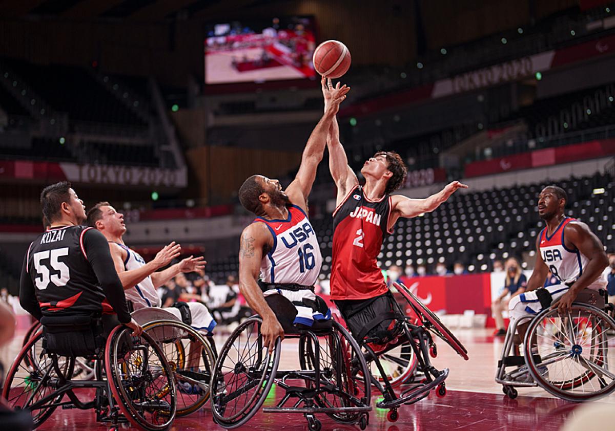 Two wheelchair basketball male athletes reach for the ball in the air