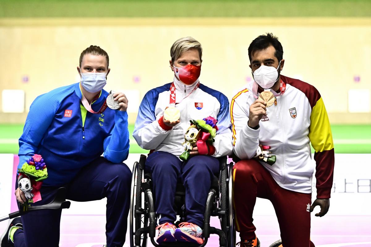 A woman and a man kneeling with their medals around a woman in a wheelchair showing her medal