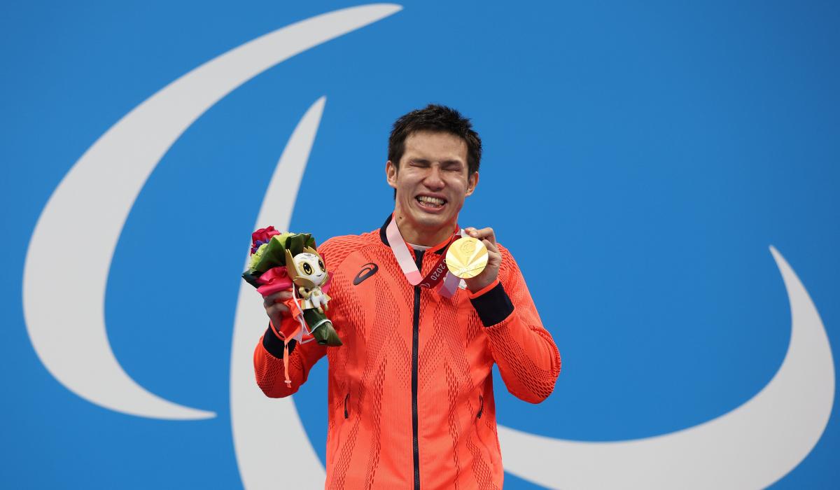 A vision-impaired athlete posing with a gold medal in left hand a mascot in his right hand