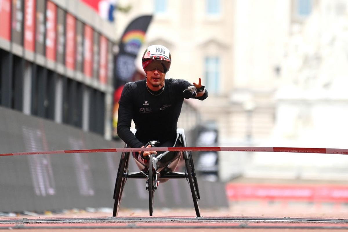 A man crosses the finish line in a wheelchair holding his left hand up with two fingers up signalizing victory