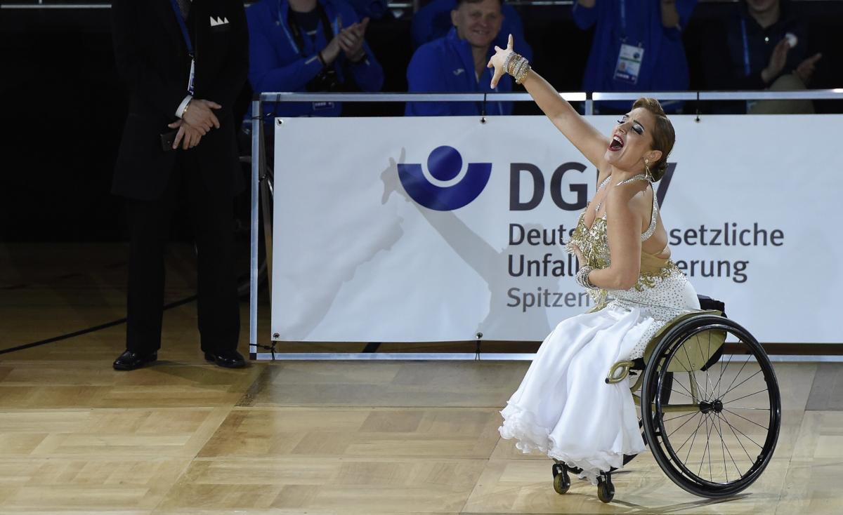 A woman in a white and gold dress dancing in a wheelchair with right arm raised