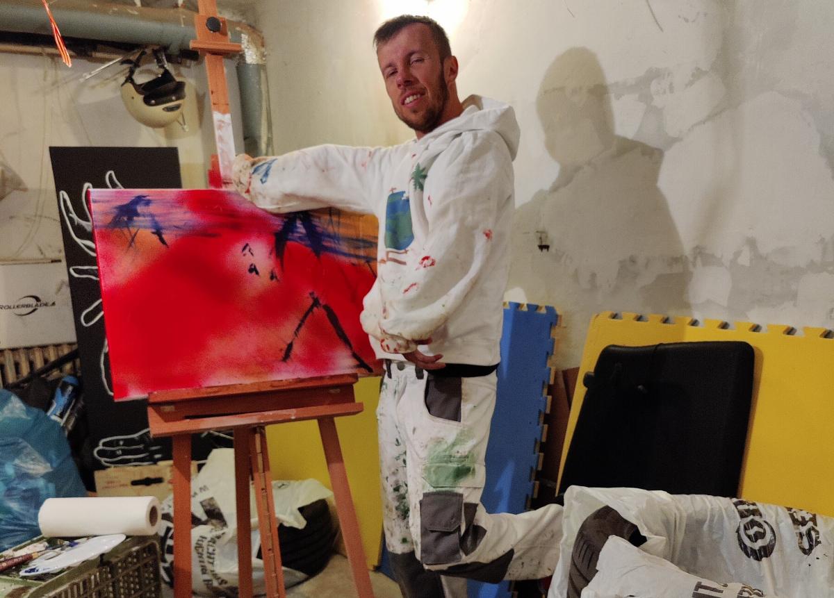 A man posing for a photo in front of his painting in a messy basement