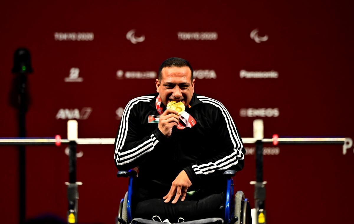 A man in a wheelchair biting the gold medal with a smile on his face