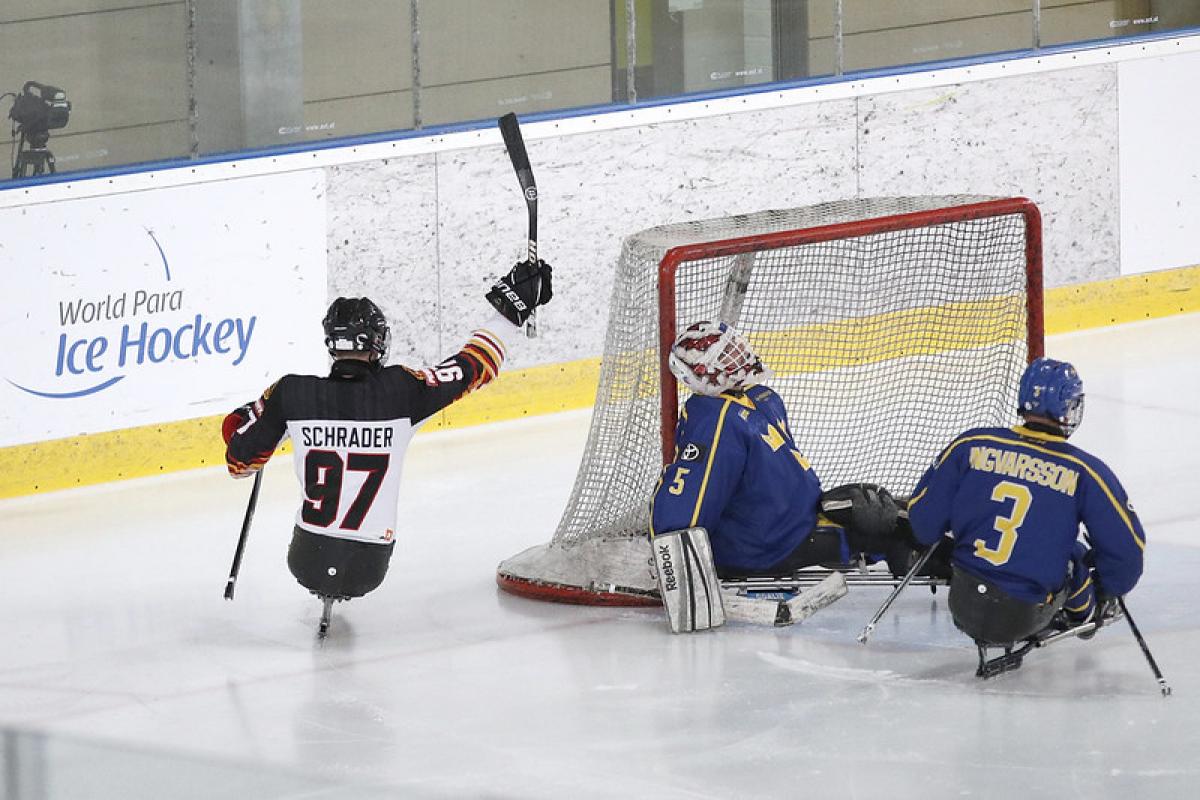 A German Para ice hockey player celebrating against two Swedish players on an ice rink