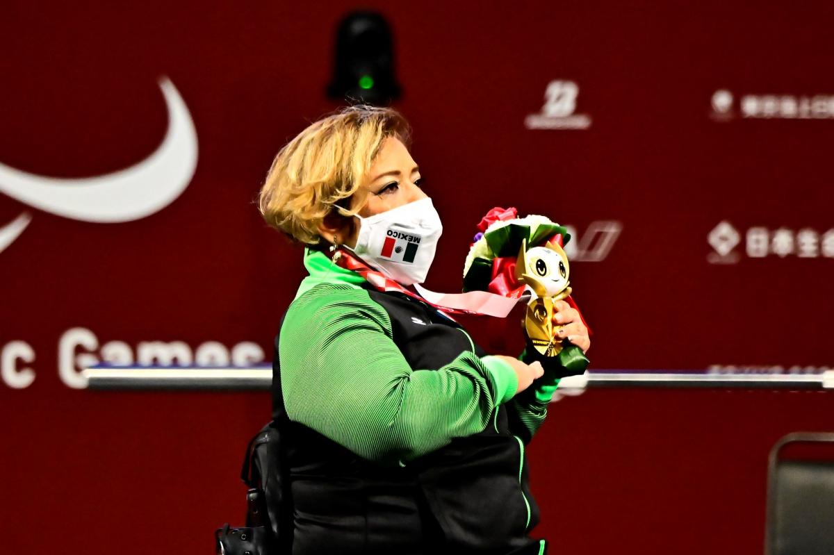 A woman in a wheelchair listening to the national anthem and holding a gold medal and a mascot in her left hand and holding her left hand high.