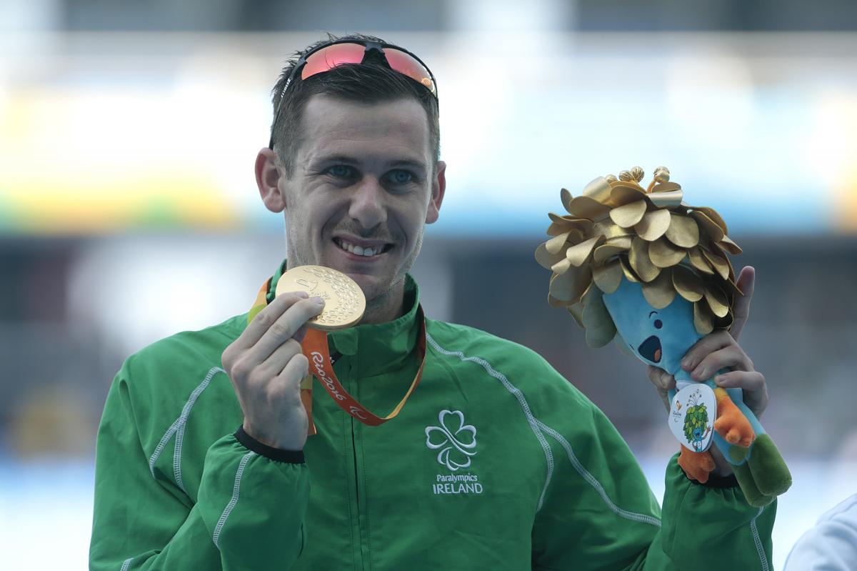 A man with the uniform of Ireland showing his gold medal and a mascot