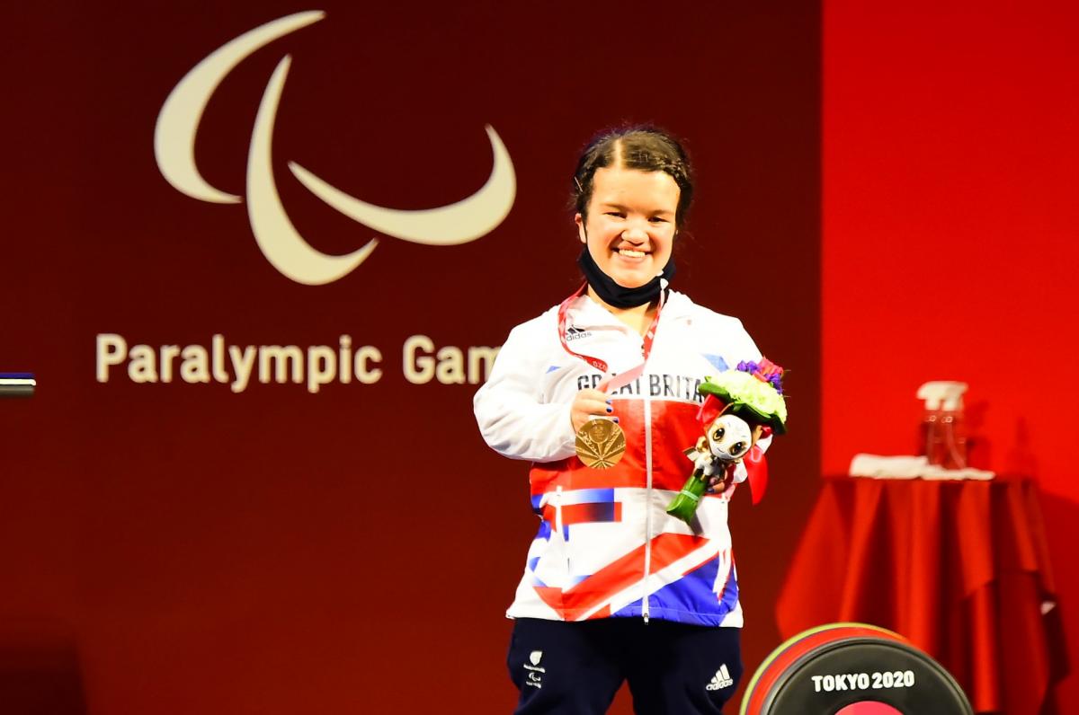 A woman holding a bronze medal in her right hand and posing for a photo on the podium.