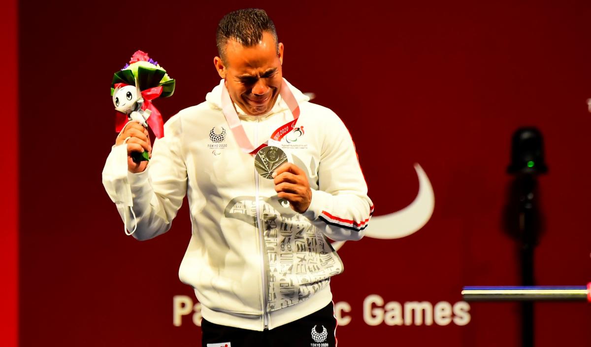 A man crying and holding a silver medal in his left hand with a mascot in his right hand.