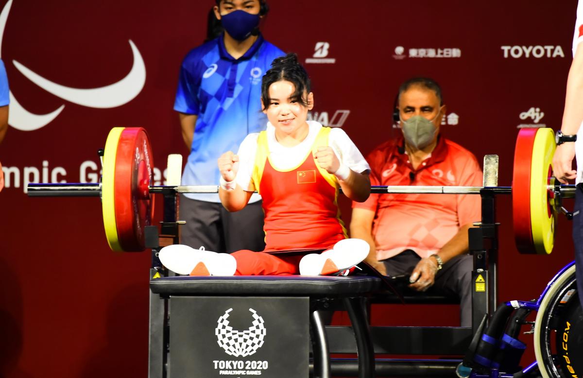 A woman sitting on a bench holding her fists high in celebration with a smile on her face