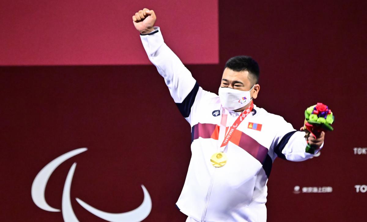 A man holding his right hand high with a gold medal around his neck and a mask on his face.