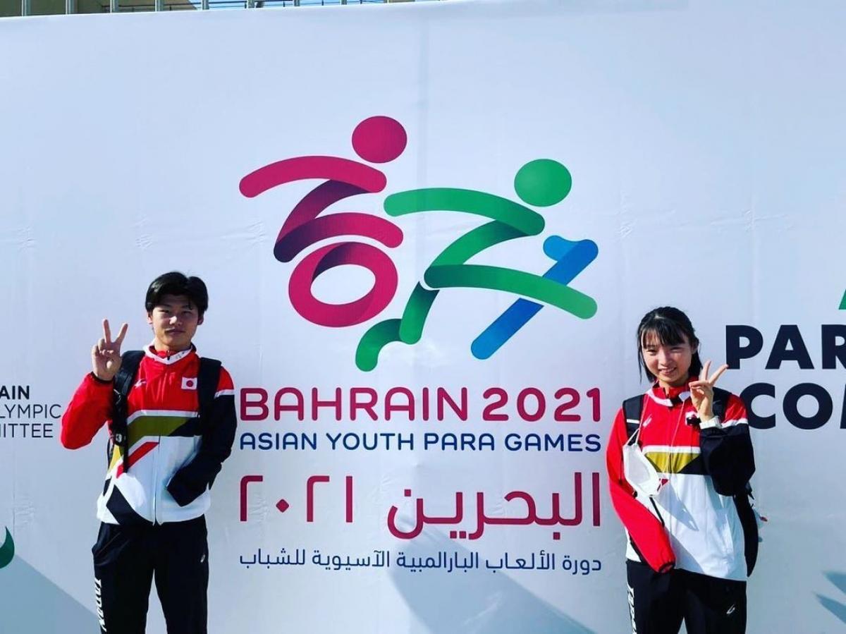 Two young Japanese Para athletes waving in front of a poster of the Asian Youth Para Games in Bahrain