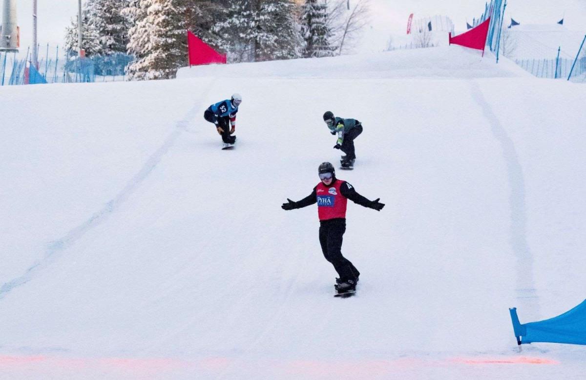 A male Para snowboarder crossing the finish line ahead of other two athletes