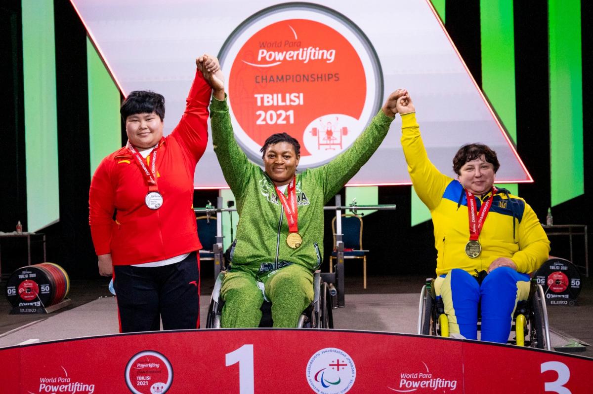 A woman in a wheelchair holding arms with a standing woman and another woman in a wheelchair in a podium