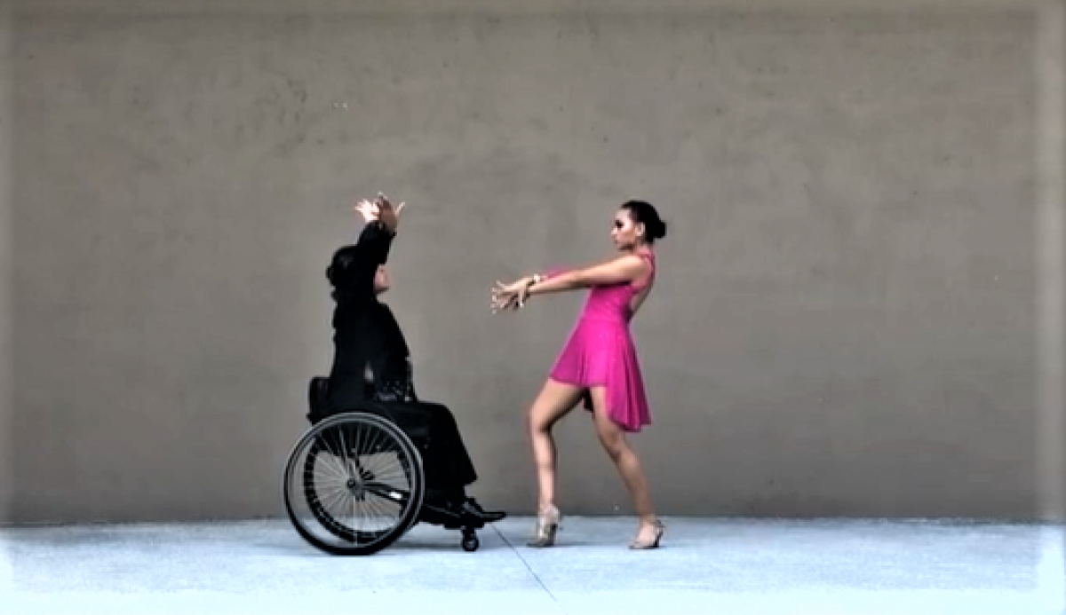 A male wheelchair dancer dressed in black dancing together with a standing woman in a pink dress