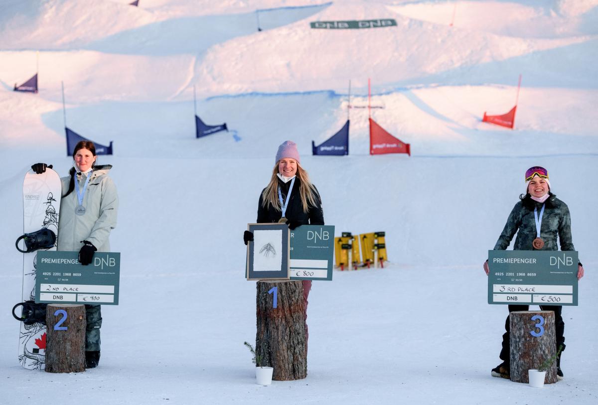 Three women smiling and showing checks in a snowboard medal ceremony in the snow
