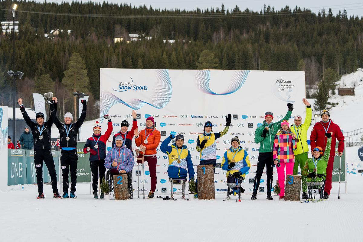 A group of 10 standing persons and four persons in wheelchairs in a medal ceremony on the snow
