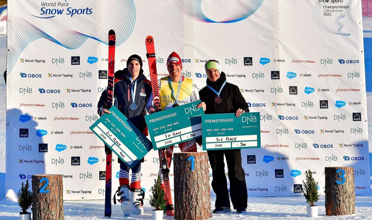 Three athletes standing in the snow in front of a white banner holding money prize.