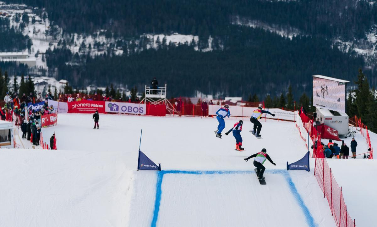 Four male athletes competing in a Para snowboard cross race. Three of them are jumping in the air, while the last one is about to jump.