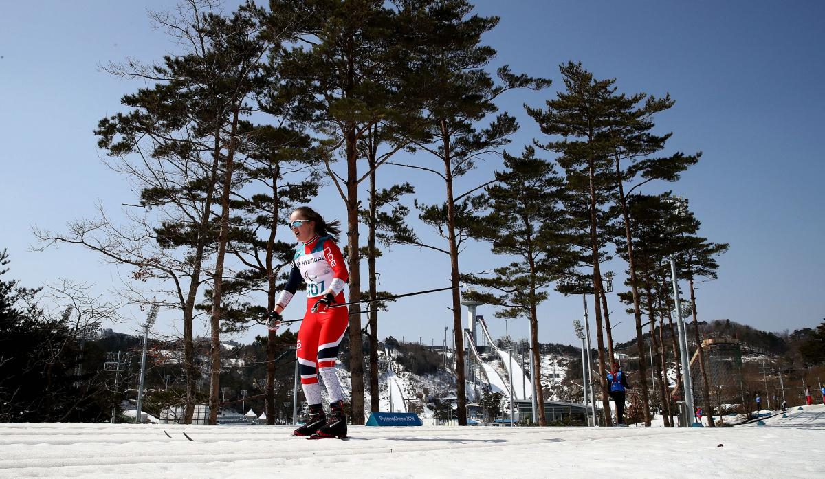 A woman in a cross-country race with goggles on her head during a sunny day. Trees are in the background with the clear and blue sky.