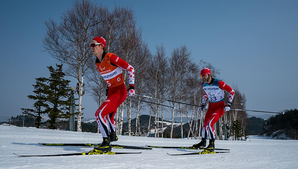 A male cross-country skier behind his male guide in a Para cross-country skiing race