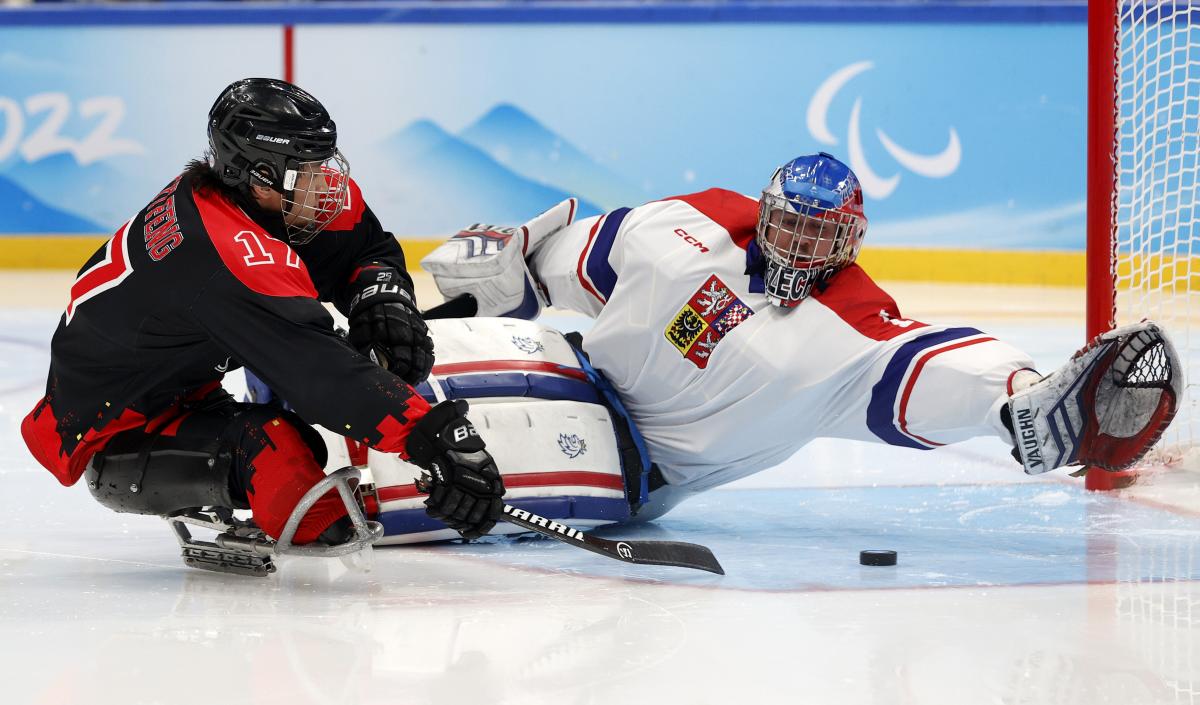 A Para ice hockey player in full gear kicks the puck with a stick past the goalie who throws himself on the ice.