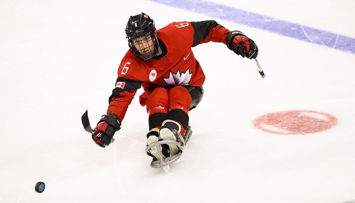 A male player on a sledge kicks the puck with a stick.