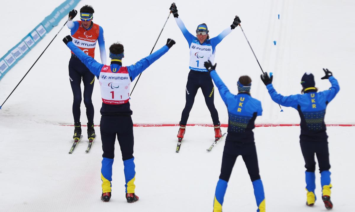 A woman crosses the finish line with raised hands and two sticks in them as the team cheers her on.