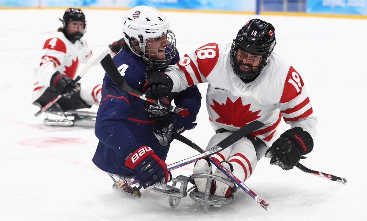 Two Para ice hockey players in full gear, one in white and the other in the blue jersey, pushing each other to gain possession of the puck.