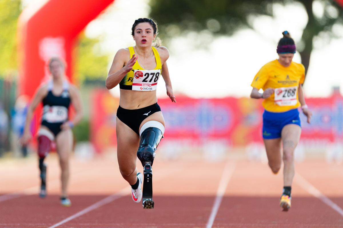 A female athlete with her left leg amputated above the knee running on the track in front of two other female athletes.