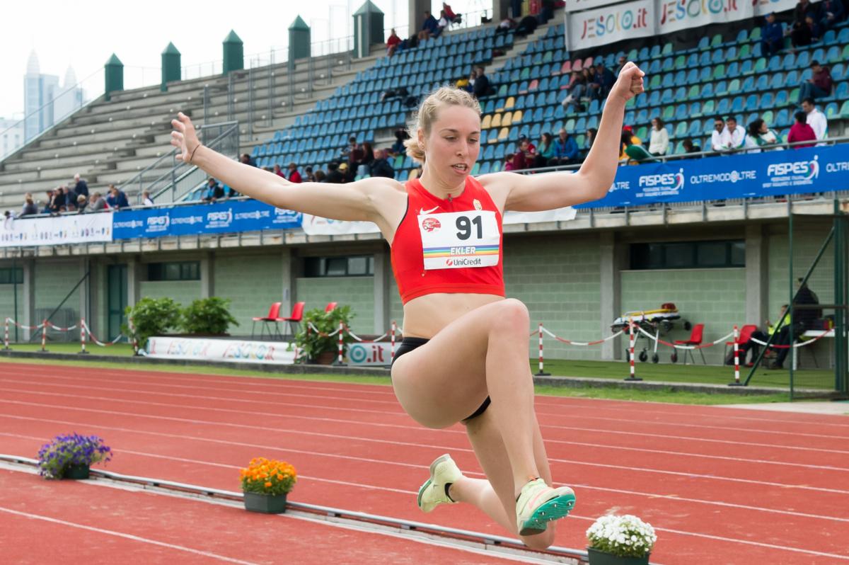 A woman in a red top jumping at the stadium with both of her hands in the air.