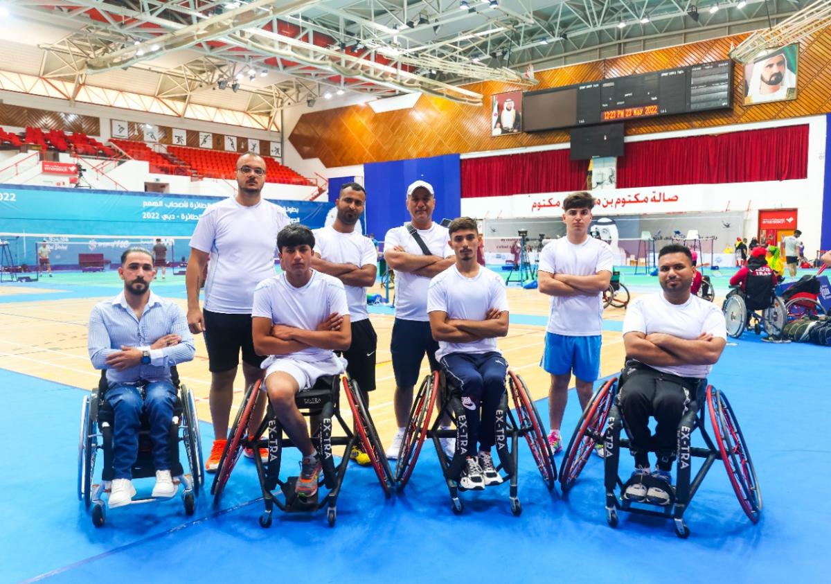 Eight athletes and staff of the Iraqi Para badminton team pose for a photo near the court.