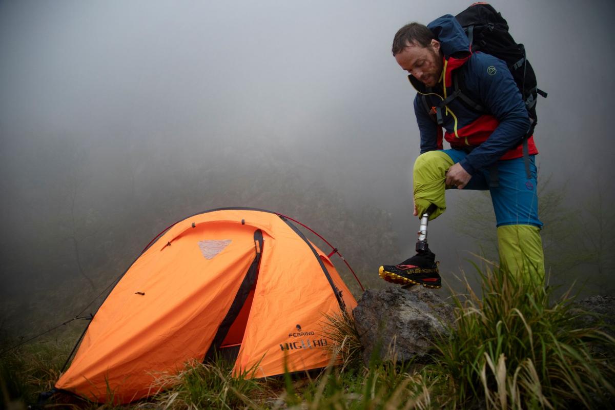 Andrea Lanfri steps on a rock as he adjusts his right prosthetic leg, with a bright orange tent on his right and heavy fog surrounding him.