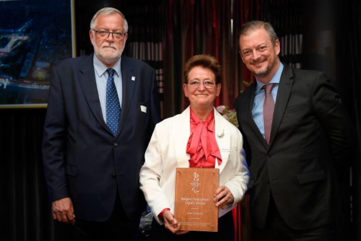 Two men in suits stand on both sides of a woman holding an awards plaque.