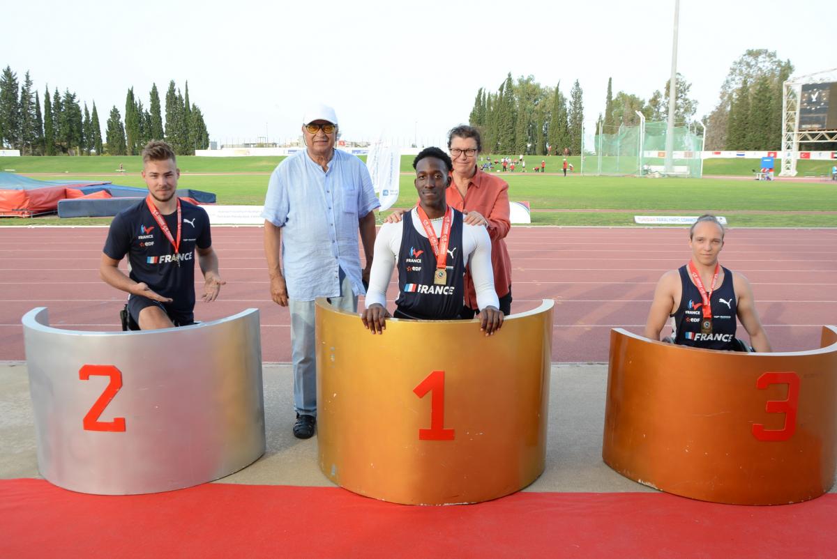 Three men on a podium with a man and a woman in the background on an athletics track