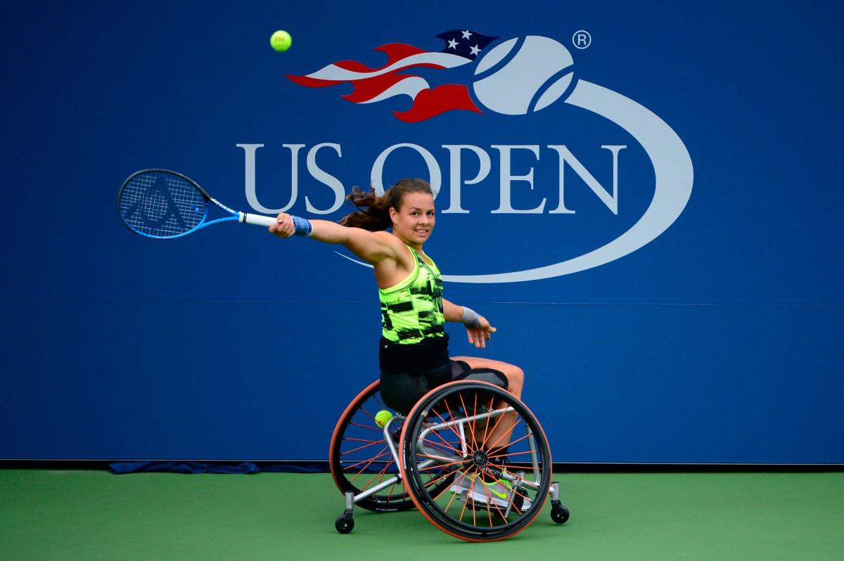 A female wheelchair player returns a shot with the words US Open written prominently on an official backdrop behind her.