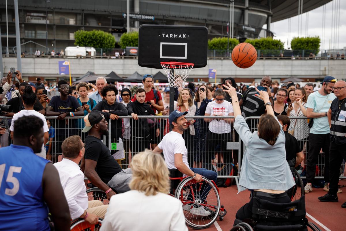 A woman sits in a wheelchair as she makes a basketball shots while a crowd of spectators looks on.