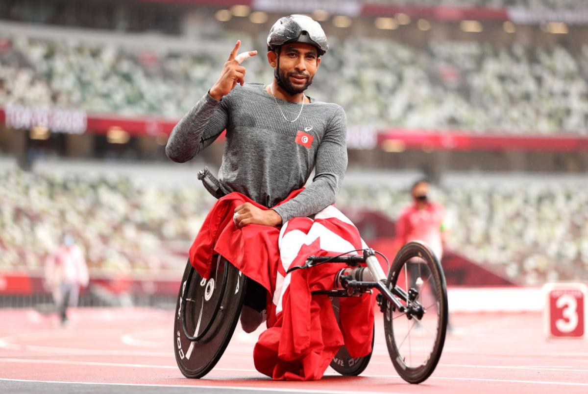 A wheelchair racer, his race chair draped in a Tunisian flag, lifts his hand in celebration on the track.