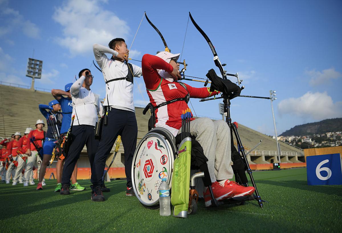 A female archer in a wheelchair shoots an arrow in a row of able-bodied archers.