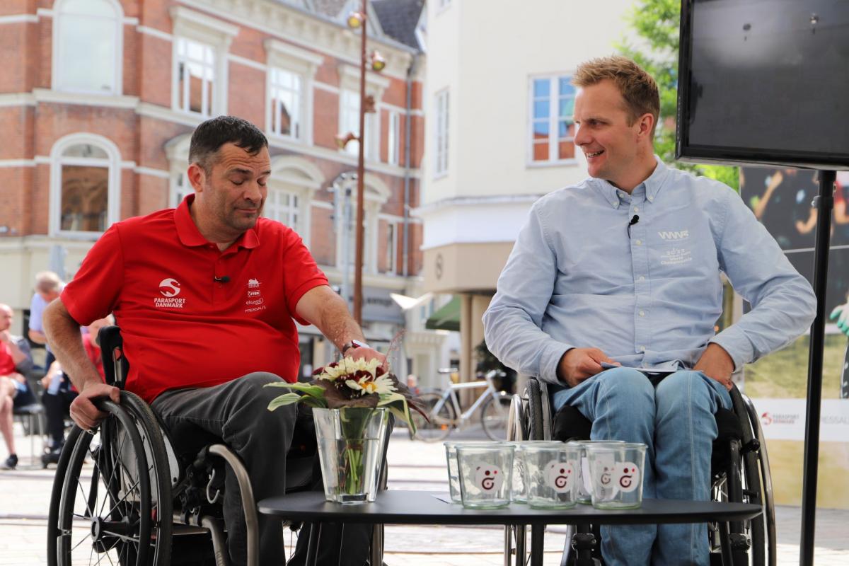Two men in wheelchairs face a table set with glasses for the 2022 Wheelchair Rugby World Championship draw.