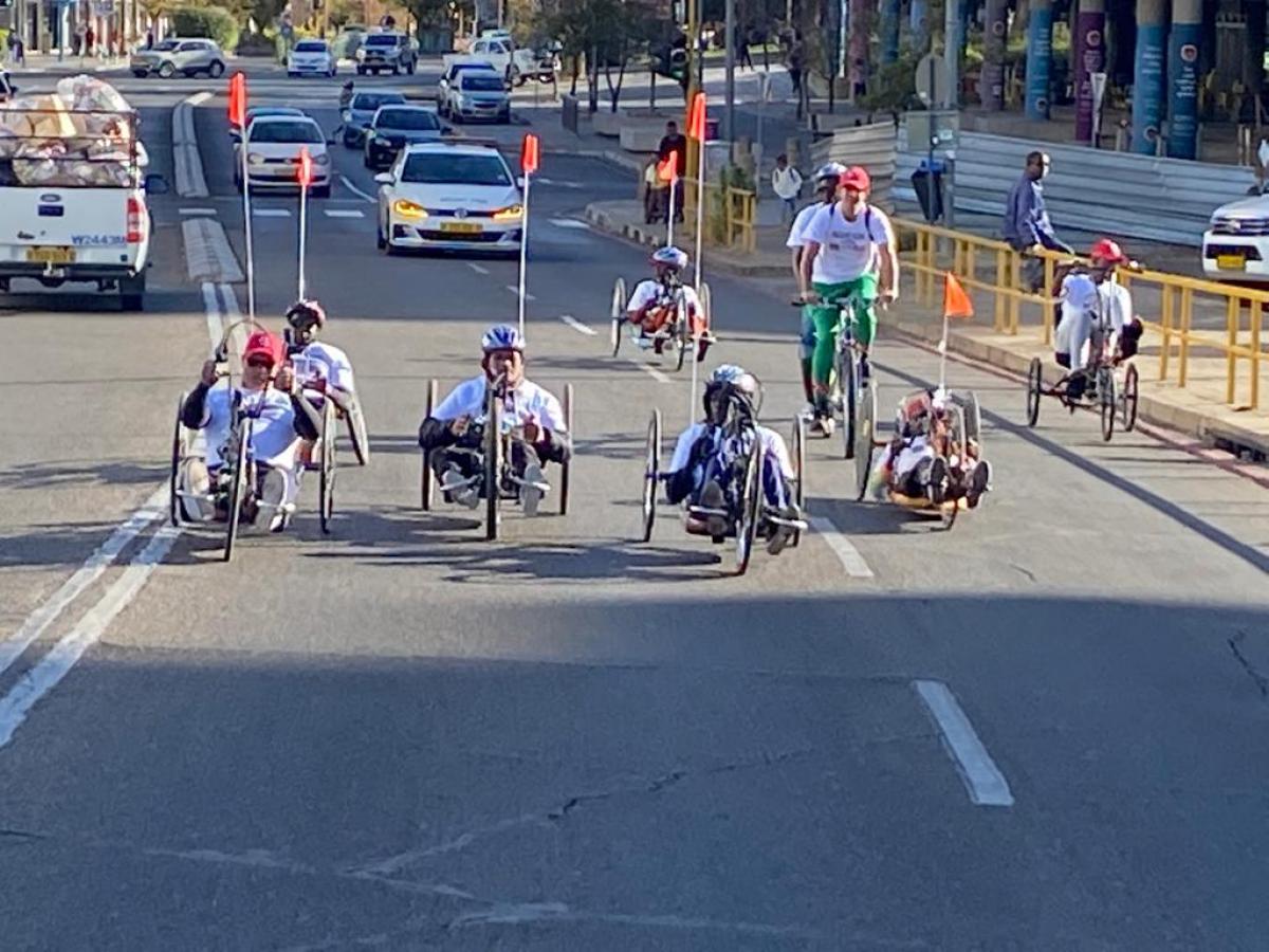 Six handcyclists ride through the city centre in Windhoek with cars following behind.