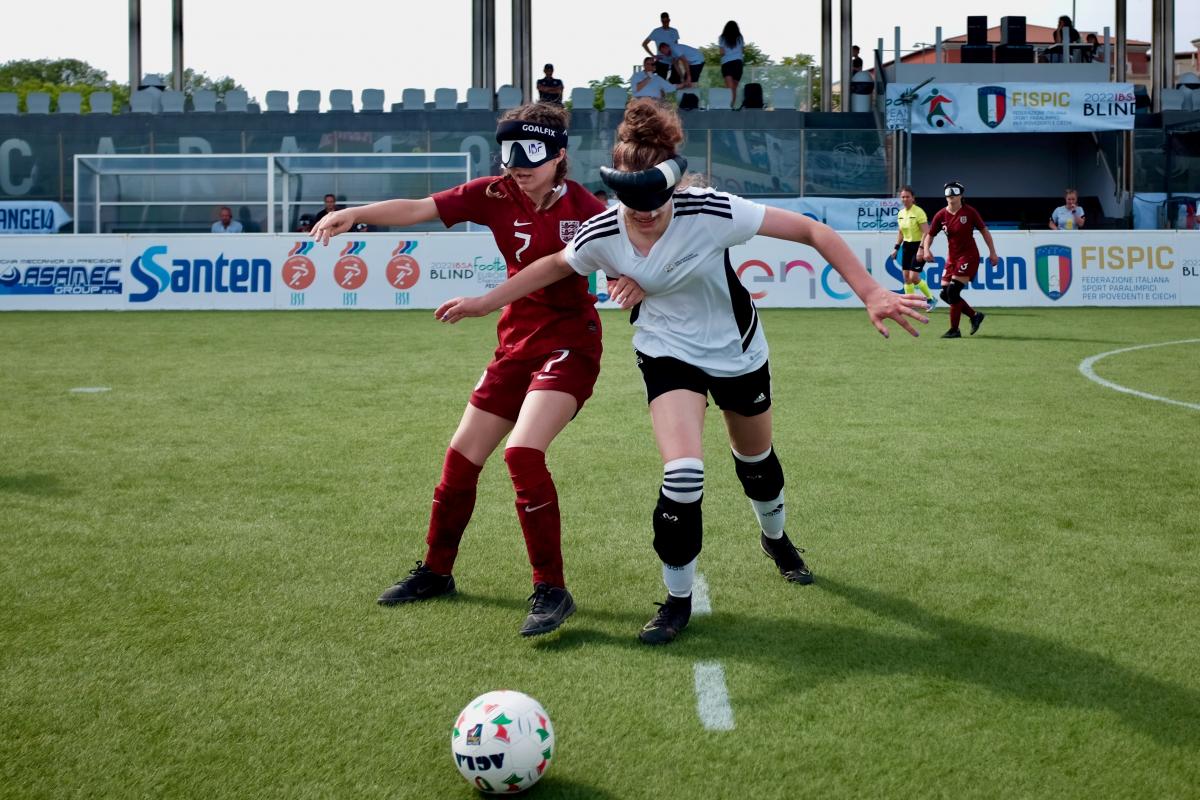 Two female players from different teams chase a ball across a football pitch.