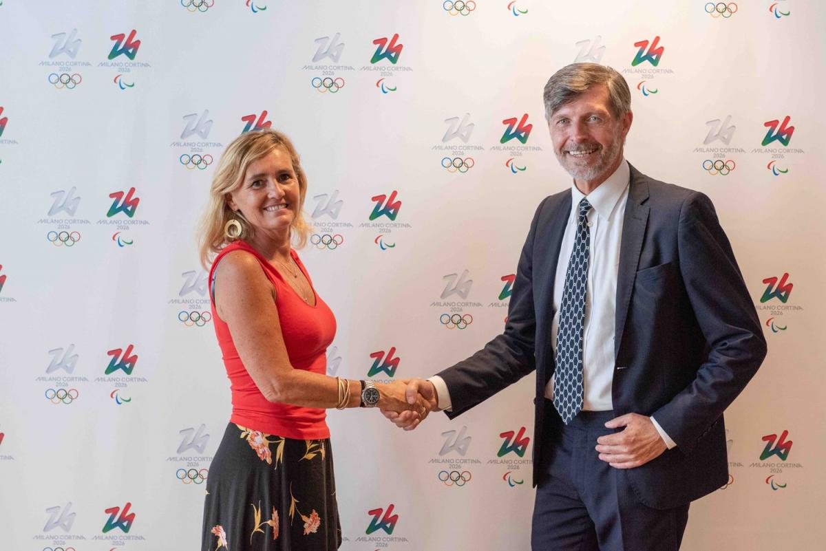 A woman and a man shake hands in front of a Milan Cortina 2026 media backdrop