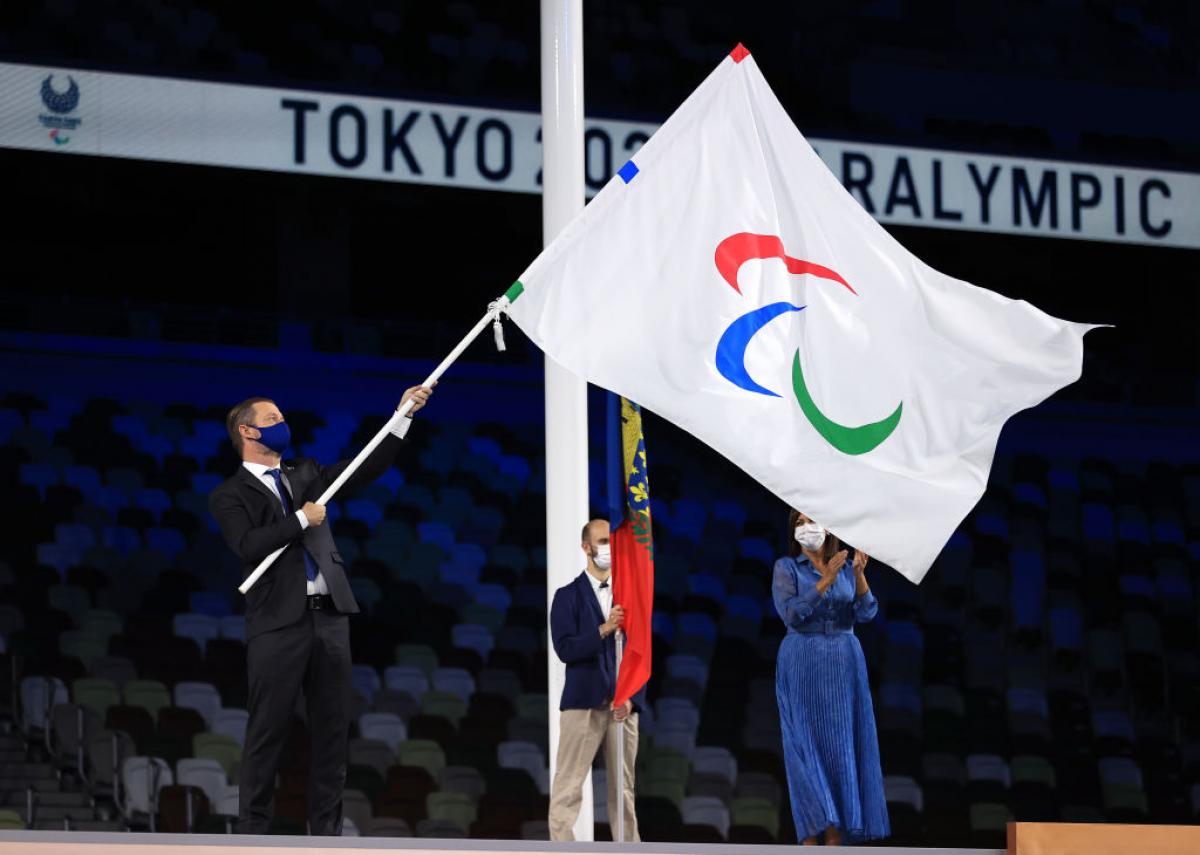 A man waves a flag with the Agitos logo on a stage at a stadium as a man and woman look on.