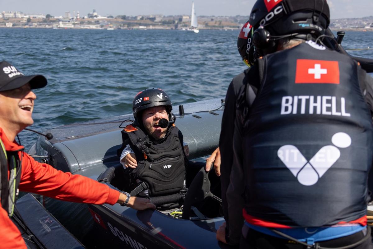 A man in a lifejacket smiles widely inside a sailboat.