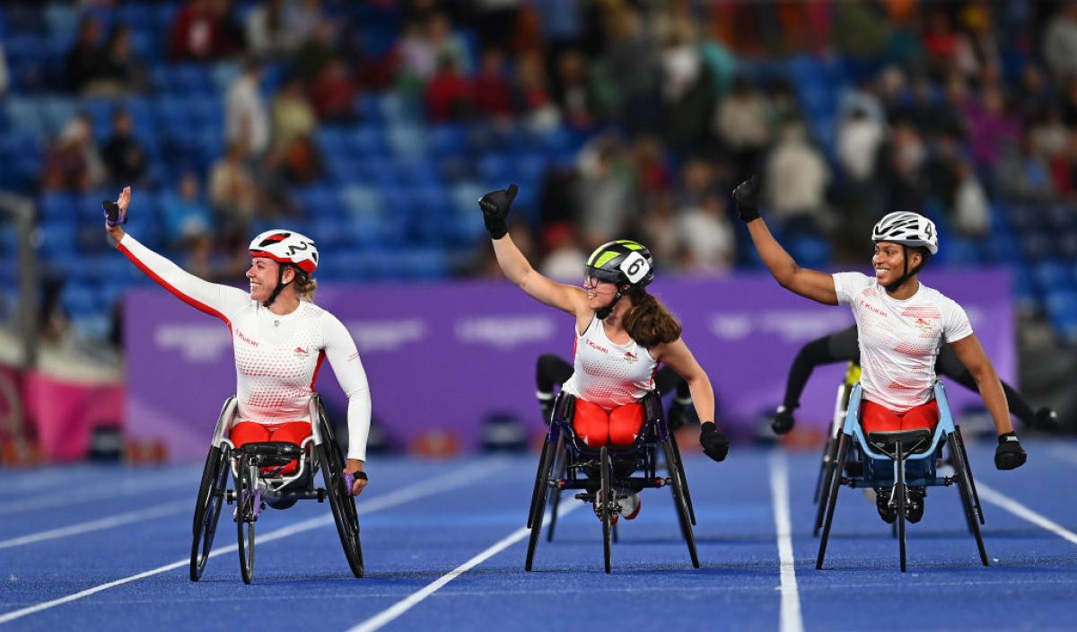 Three female wheelchair racers on a blue track in a crowded stadium